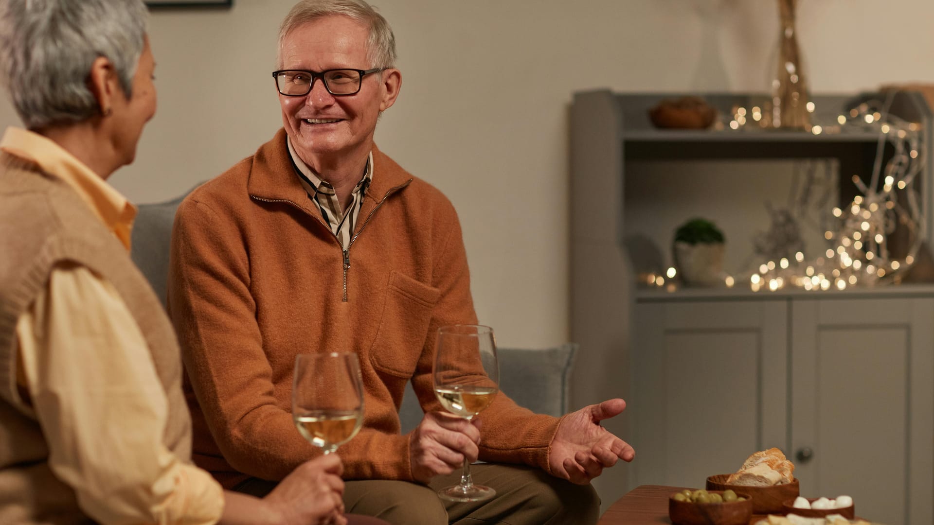 A smiling senior man in an orange sweater holding a glass of wine while talking with a friend