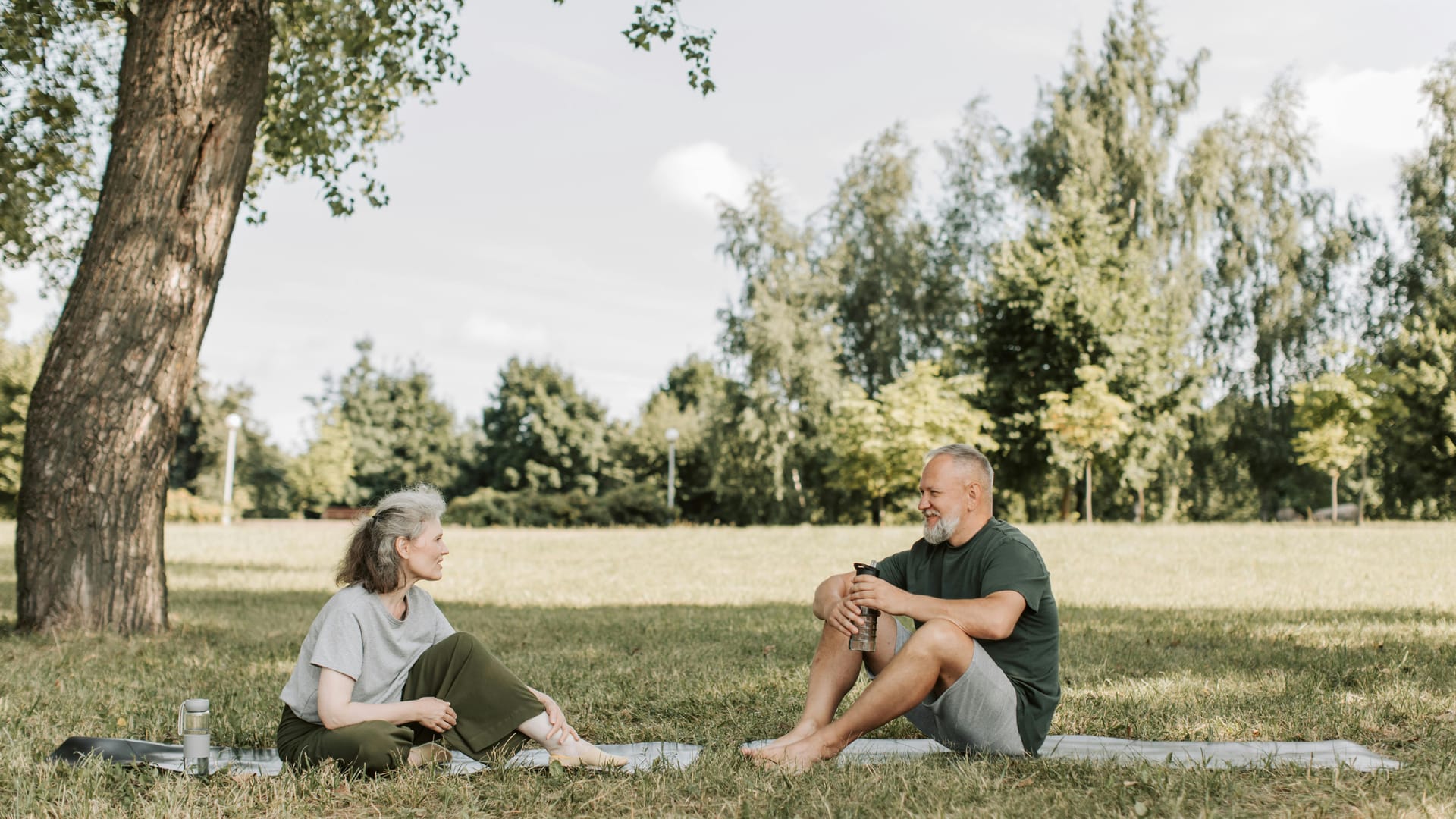 A senior couple sitting on yoga mats in a park, talking under the trees