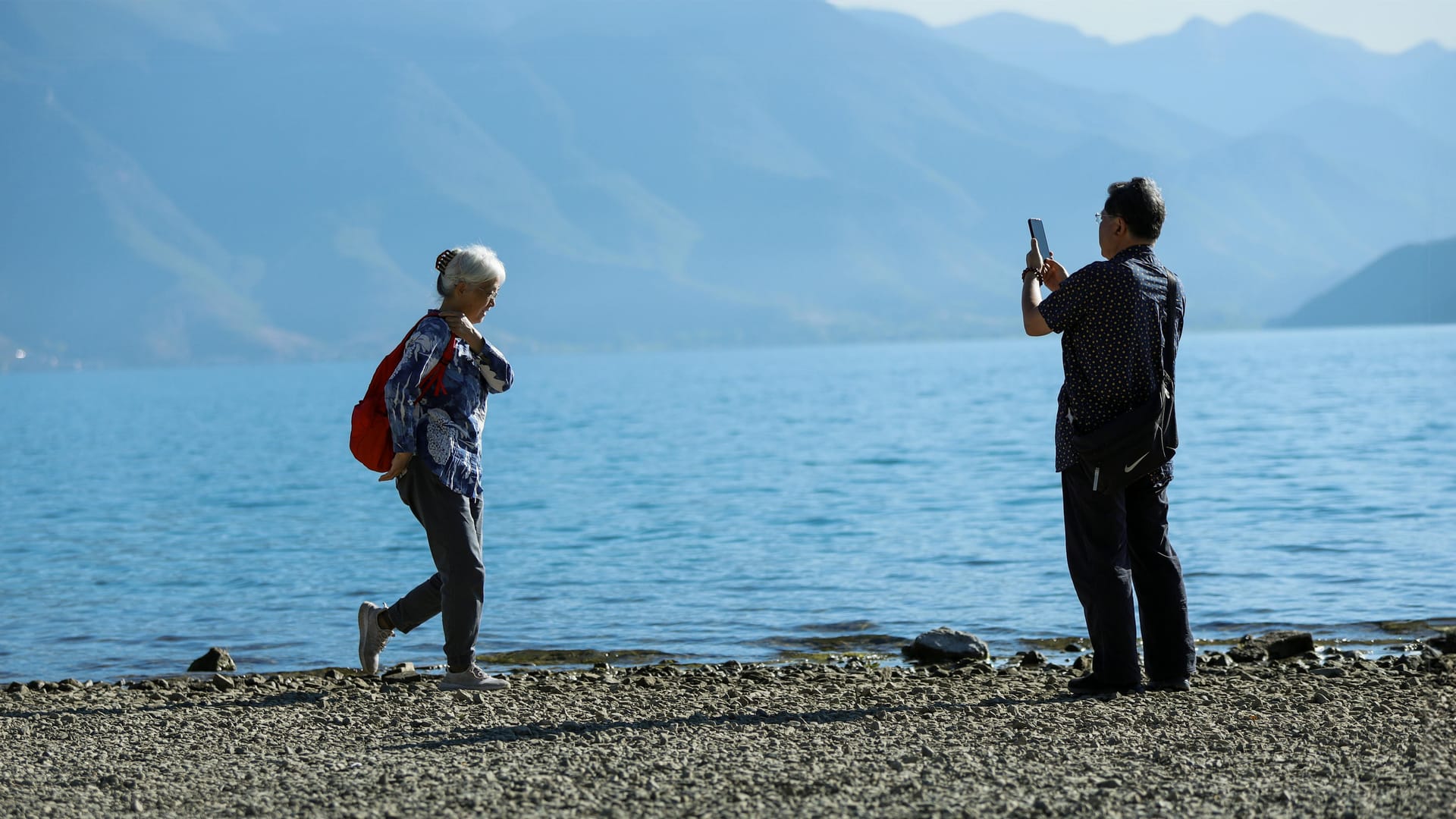 A senior couple on a lakeshore taking a photo, with mountains behind them